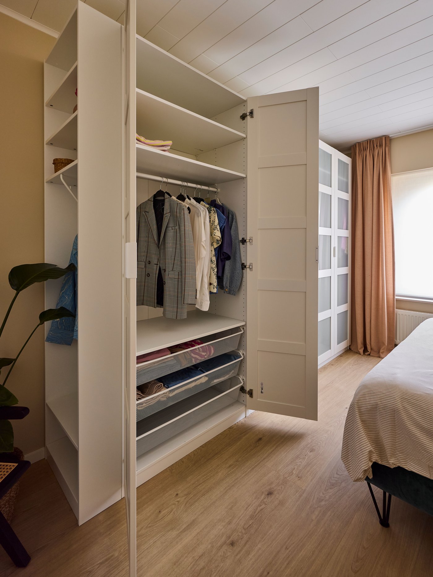 Open kast met hangende kledij, planken en lades in een slaapkamer naast een bed en gordijnen.
