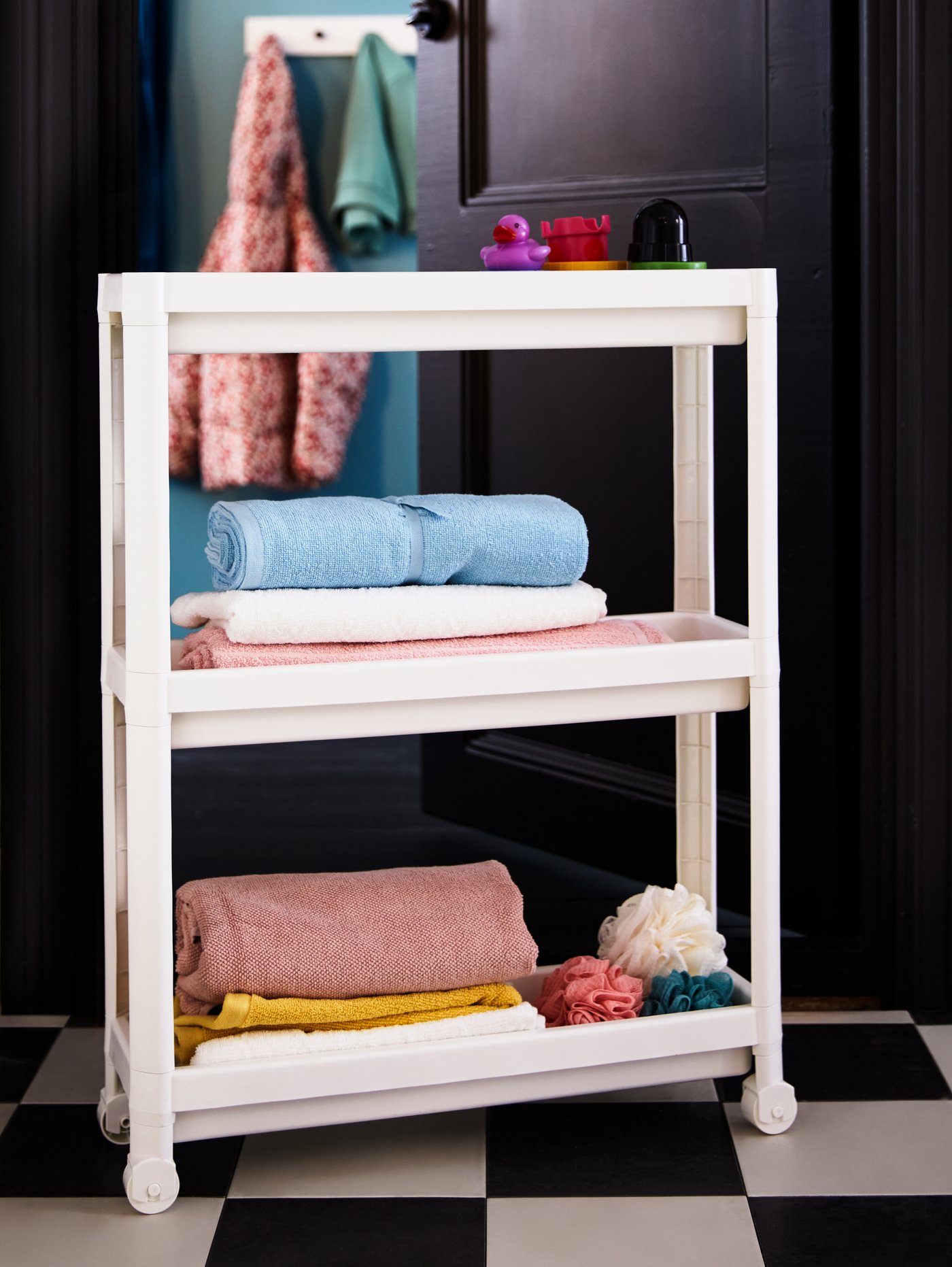 A bathroom with white and black floor tiles and a white VESKEN shelf unit with body puffs and towels in pastel colours.