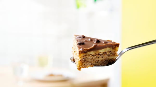 Vork met een stuk romige laagjestaart met chocoladeglazuur op de voorgrond, tegen een zachte, lichte achtergrond met onscherpe details.