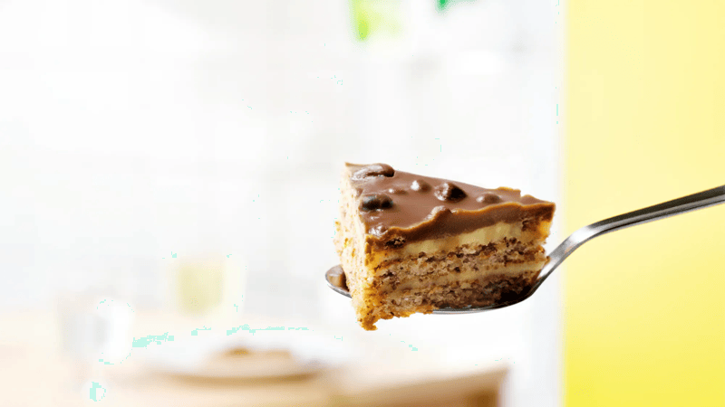 Vork met een stuk romige laagjestaart met chocoladeglazuur op de voorgrond, tegen een zachte, lichte achtergrond met onscherpe details.