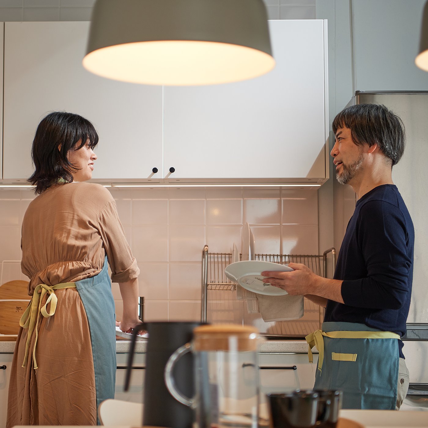 明るいキッチンで2人が食器を洗う様子。白いキャビネットとステンレス冷蔵庫、木製カウンターがあるモダンな空間。