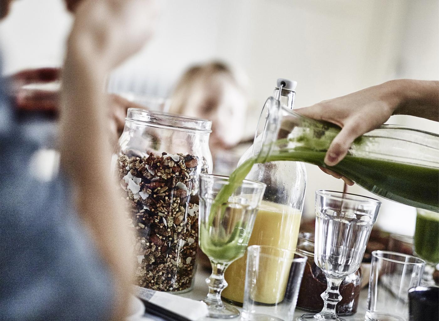Hand giesst grünen Smoothie aus einem Mixer in Glas, daneben Gläser, Orangensaft und ein grosses Glas mit Müsli.