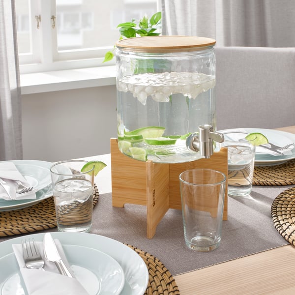Un dispenser pieno  in vetro con rubinetto IKEA 365+ con supporto in bambù al centro di una tavola apparecchiata. Sotto il rubinetto, un bicchiere. 