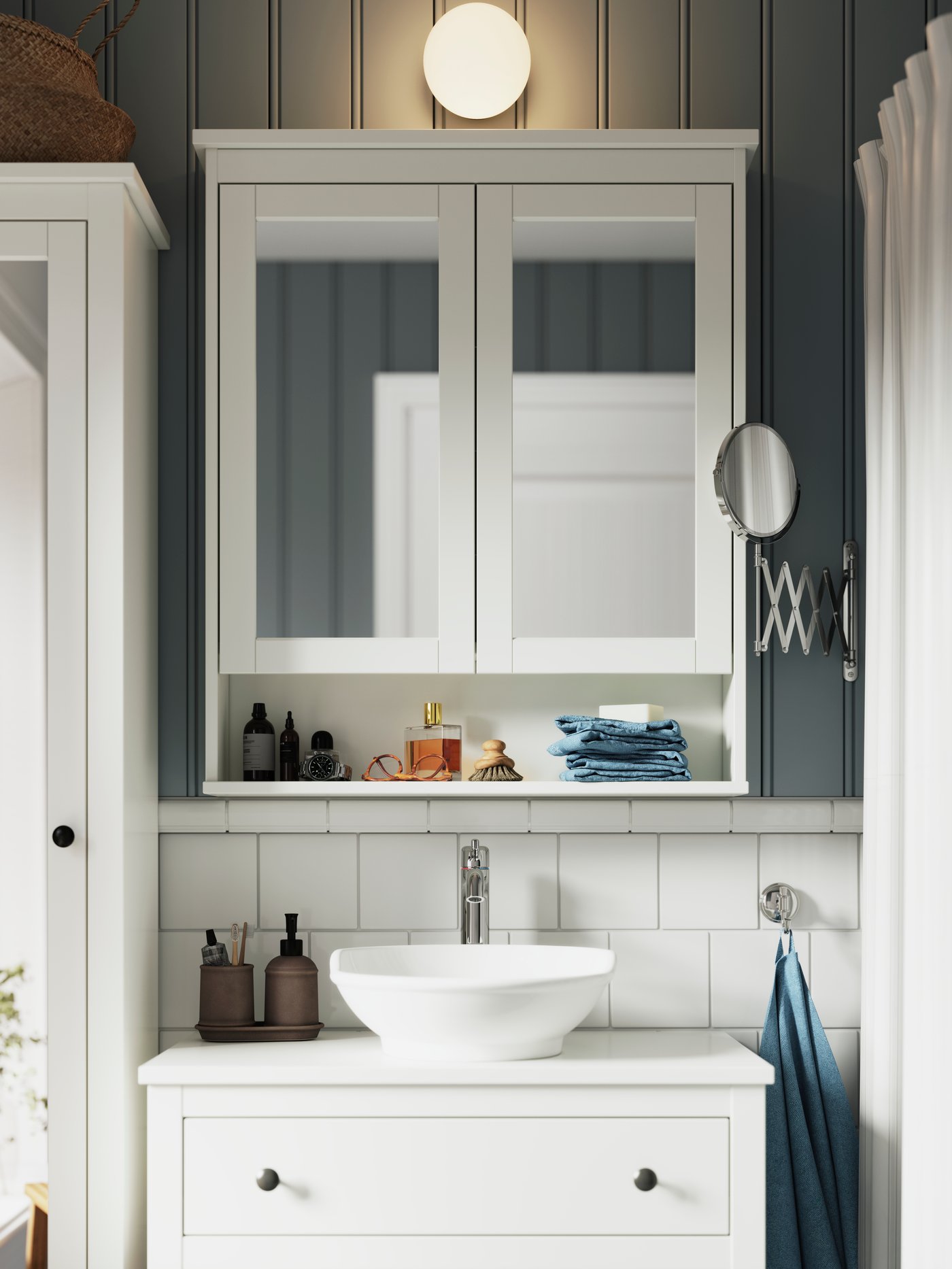 A FRIHULT wall lamp in metal and glass is mounted above a white HEMNES mirror cabinet and a white HEMNES wash-stand.