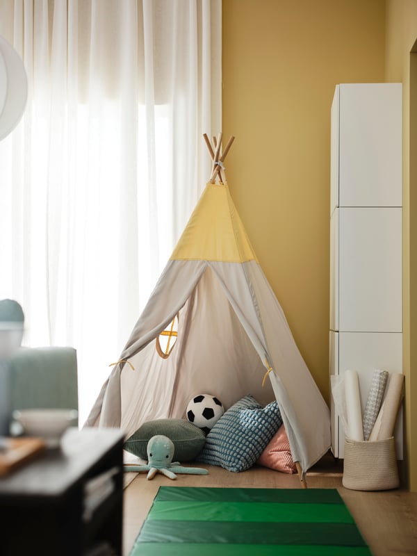 A HÖVLIG children’s tent is in the corner of a yellow living room with cushions and an octopus BLÅVINGAD soft toy in it.