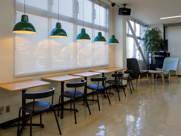 窓際に木製カウンターテーブルと黒いスツールを並べたワークスペース。緑のペンダントライトと奥に観葉植物が見える。