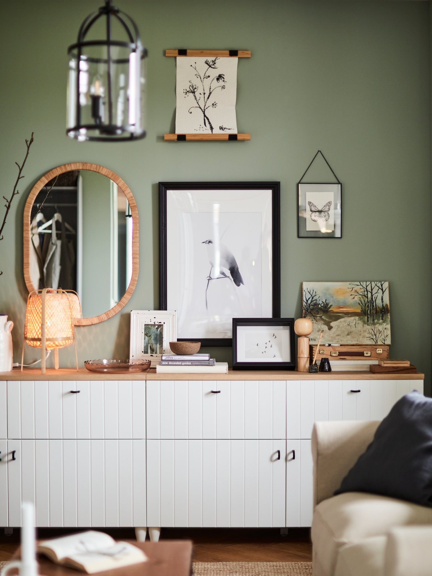 A green wall with framed drawings of a bird, flowers and a butterfly, a bamboo lamp, a mirror, a white cabinet.
