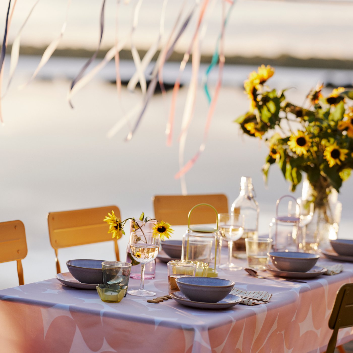 In a summery atmosphere, a table is set for dinner by the water, featuring TOFSAND tablecloth in light pink. Yellow flowers.