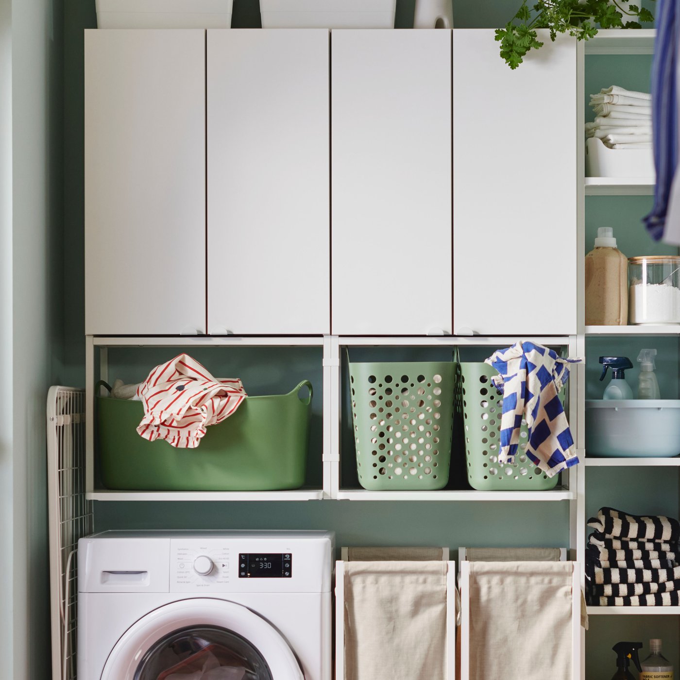 Due telai per mensole a muro ENHET bianchi affiancati con ripiani profondi contengono cesti della biancheria sopra una lavatrice, sotto gli armadietti.