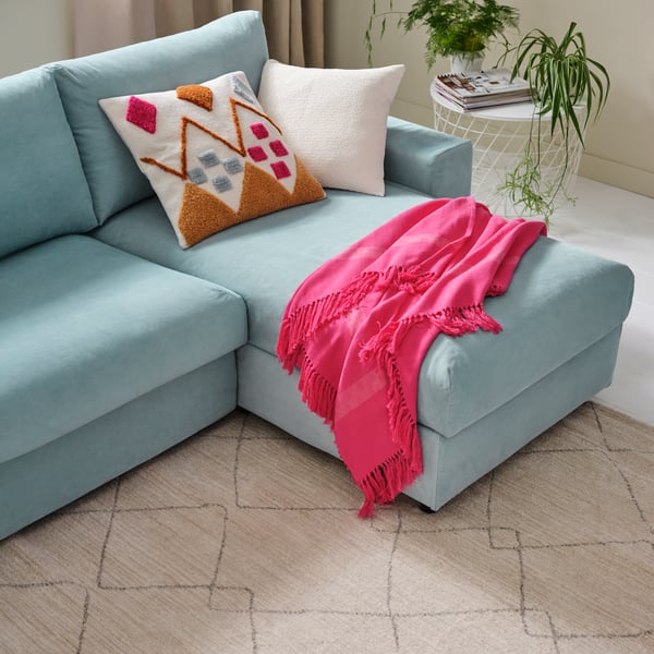 Hellblaues Sofa mit verspielten Kissen und knallpinker Decke in einem hellen Raum mit Pflanzen und Teppich mit Rautenmuster
