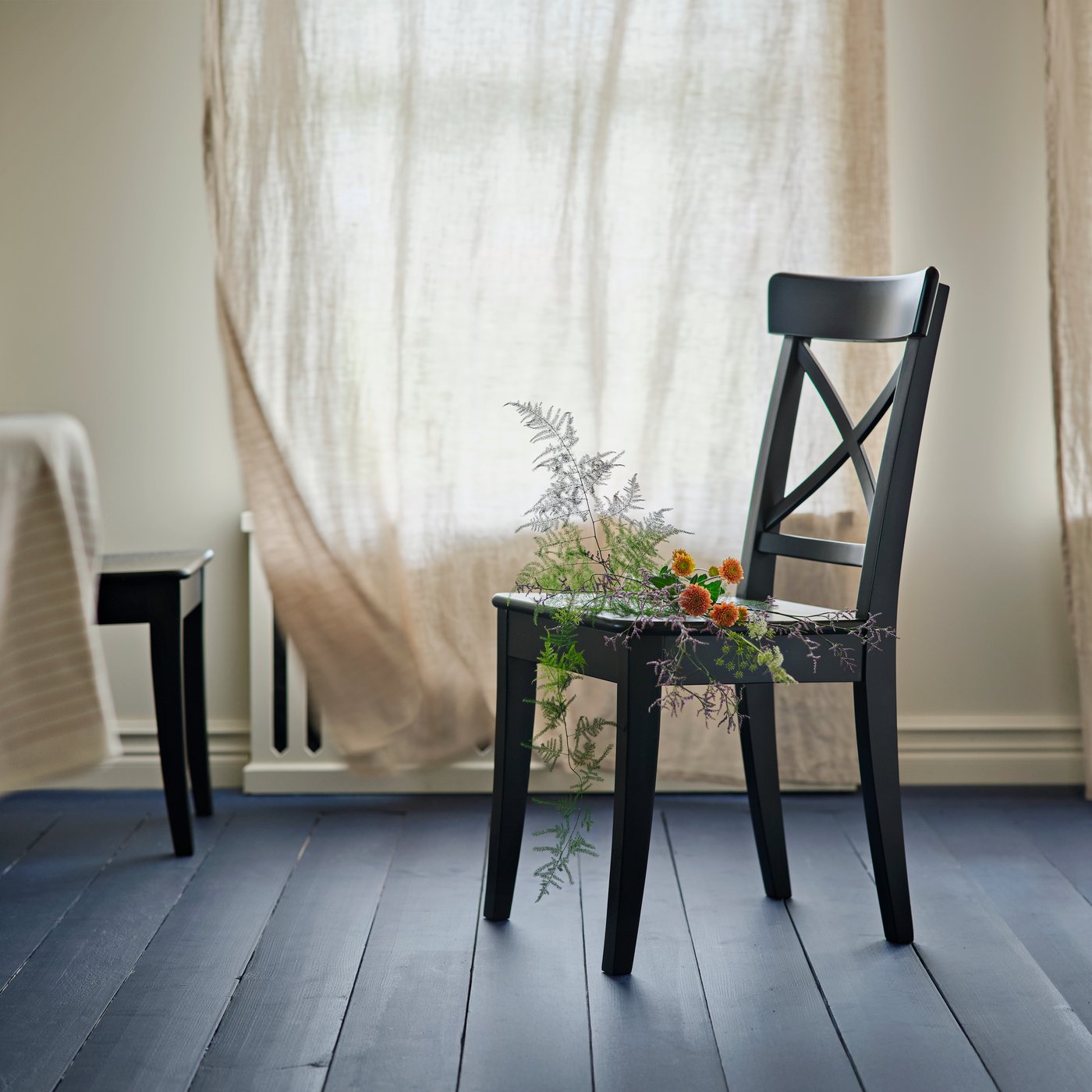 Ein schwarzer ROSENTORP Stuhl mit Grünpflanzen und orangefarbenen Blumen auf der Sitzfläche steht in einem hellen, luftigen Raum.