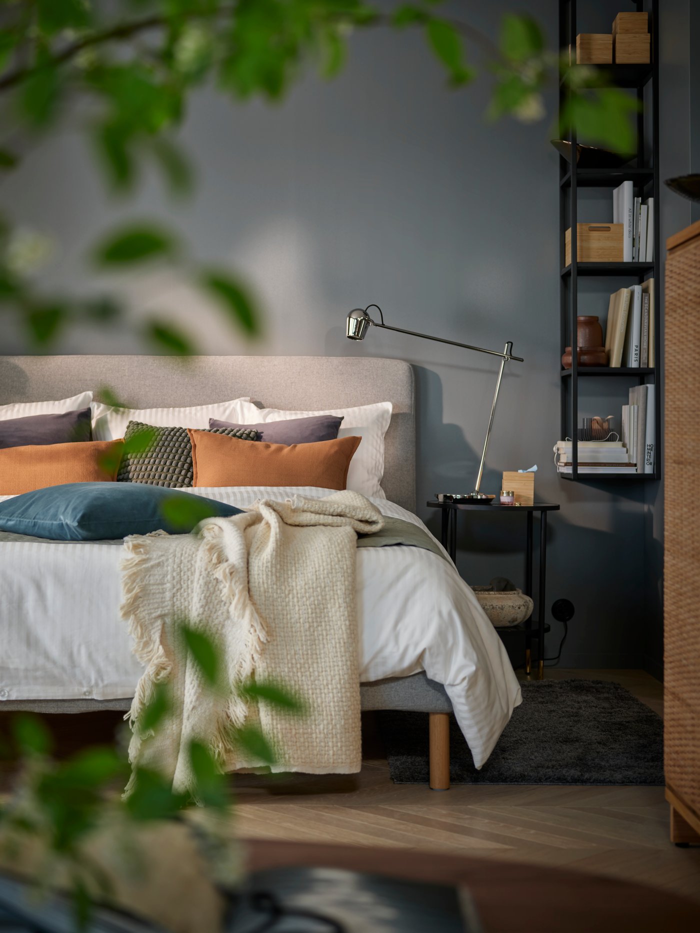 Un dormitorio con paredes grises y vista de una cama, una mesa de noche y estantes. En primer plano, hojas verdes difuminadas de una planta.