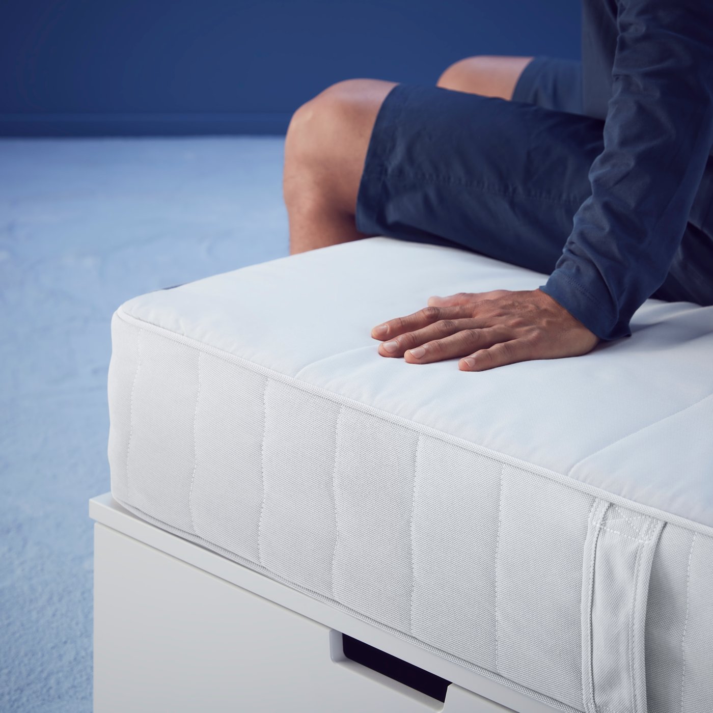 Un hombre sentado en la esquina de una cama con la mano apoyada en el colchón.