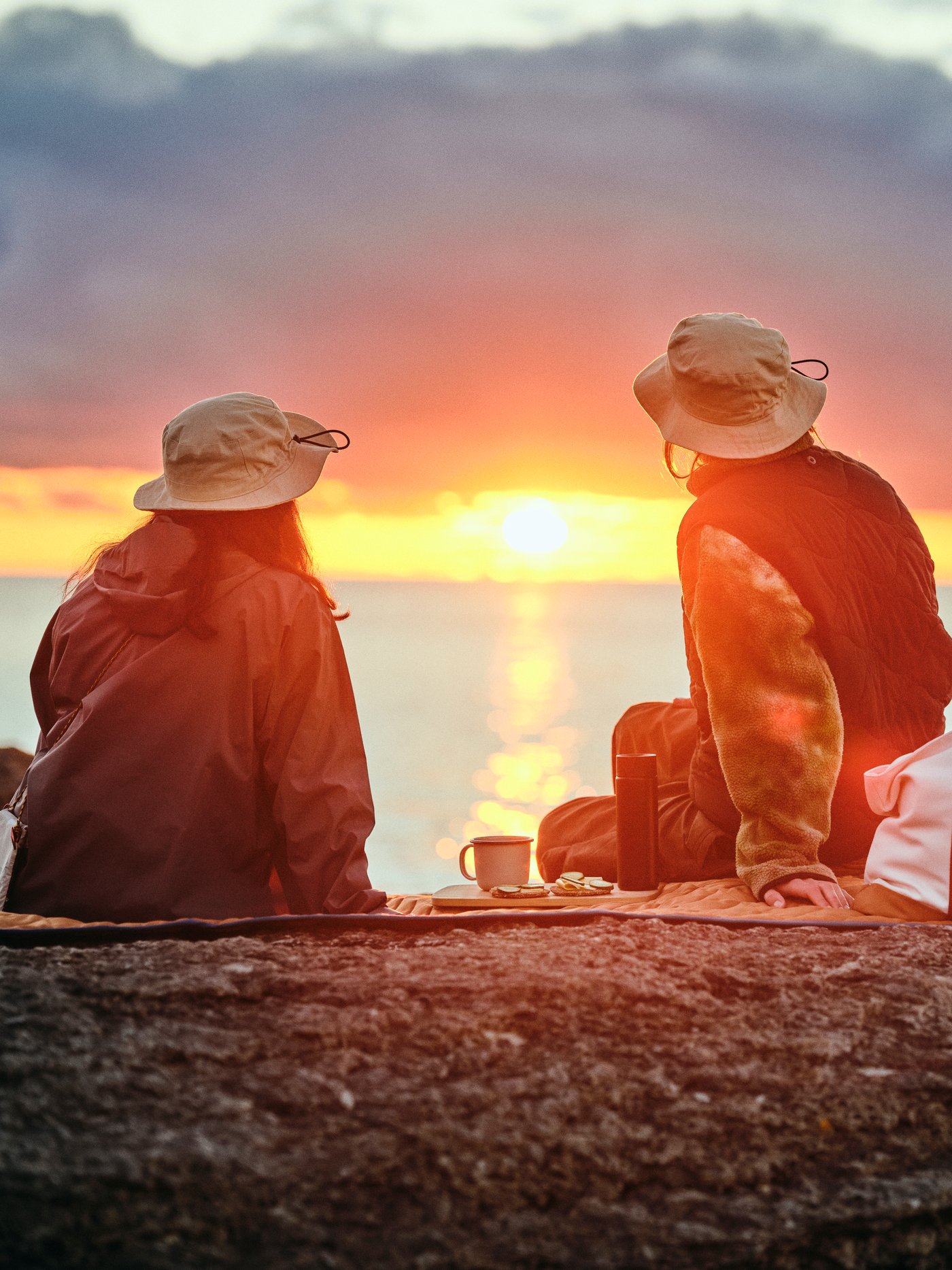 Due persone con cappelli SOLUPPGÅNG sono sedute su una coperta da picnic SOLUPPGÅNG mentre guardano il tramonto sul mare.