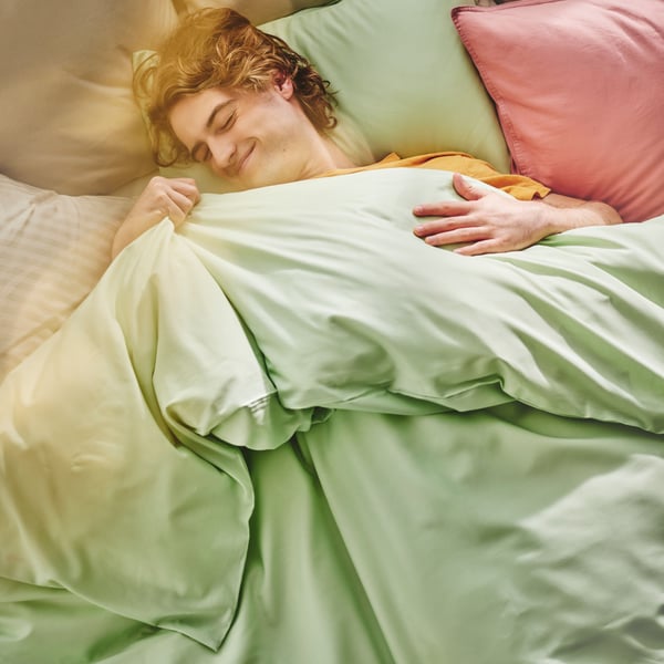 A person smiles as they lie in bed covered with light green PILTANDVINGE bed linen, with other pillows around them.