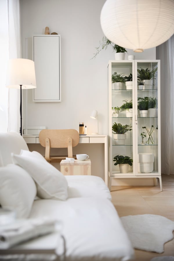 A white desk and natural wood chair create a study space in a multifunctional living room with plants in a glass cabinet.