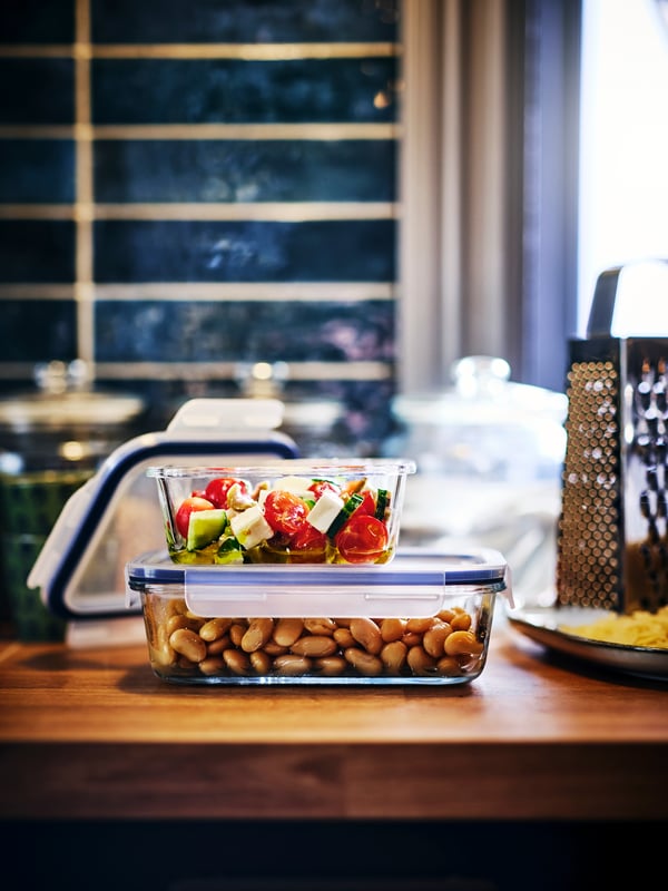 Two food-filled IKEA 365+ food containers with lids on top of one another on a wooden worktop, with a grater to the right.