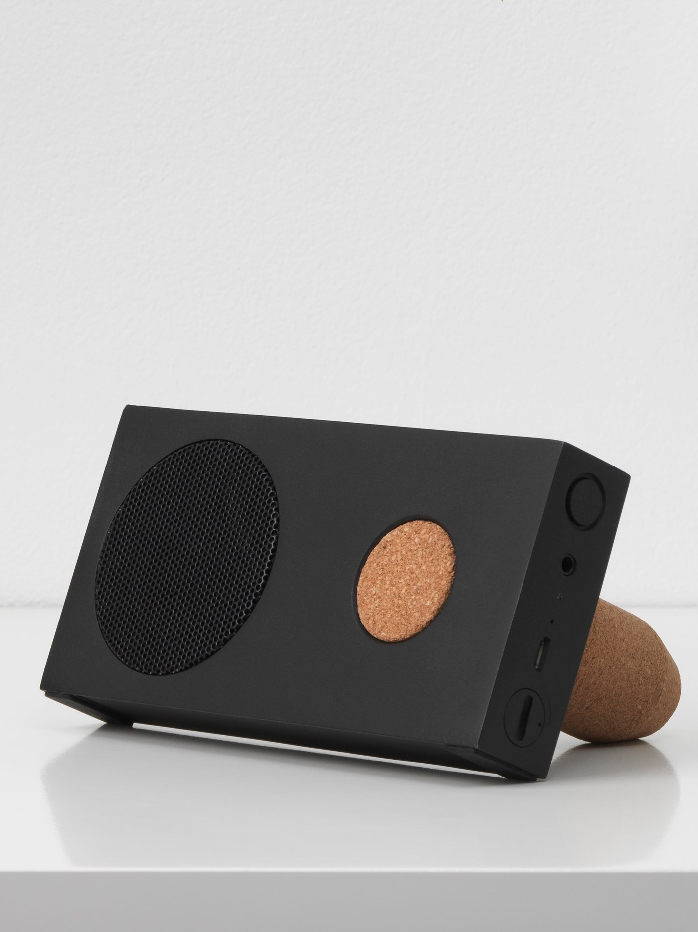 Svart bärbar ENEBY högtalare med hållare i kork står på en vit yta mot en vit vägg.