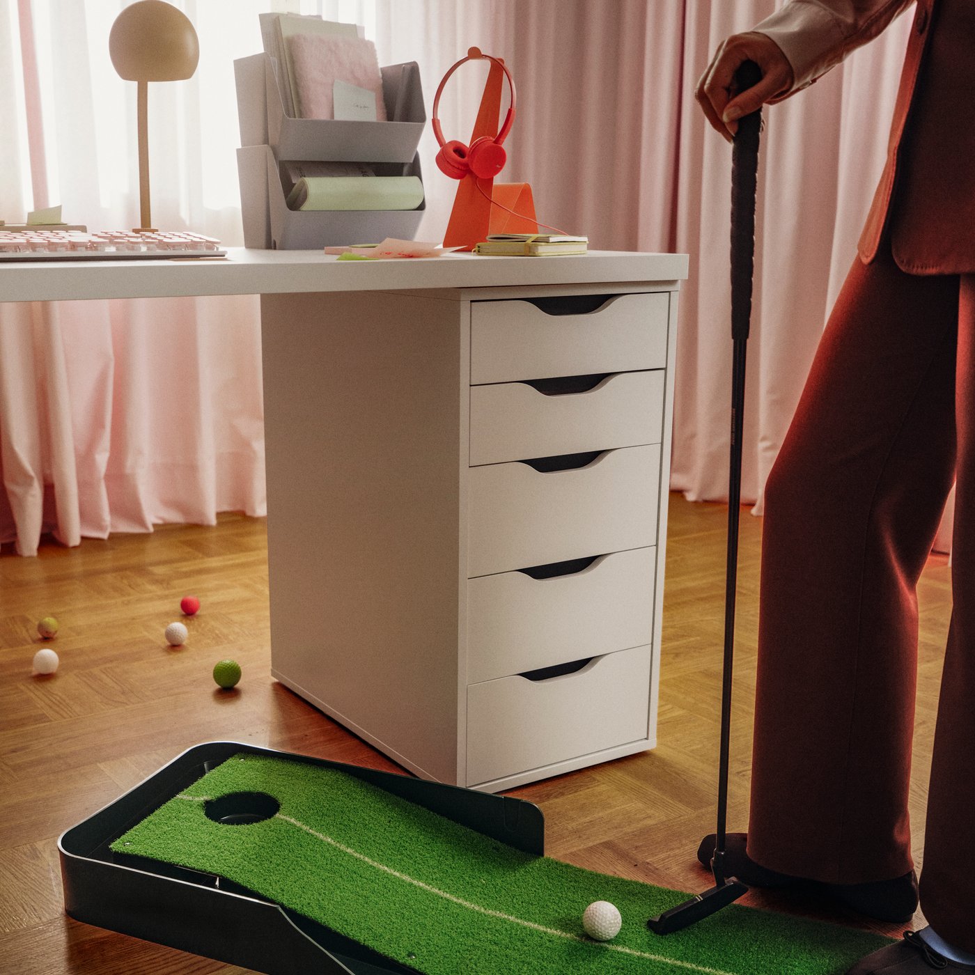 Una cassettiera ALEX bianca, dotata di eleganti maniglie a incavo, sostiene una scrivania bianca. Accanto, una persona si esercita nel minigolf.