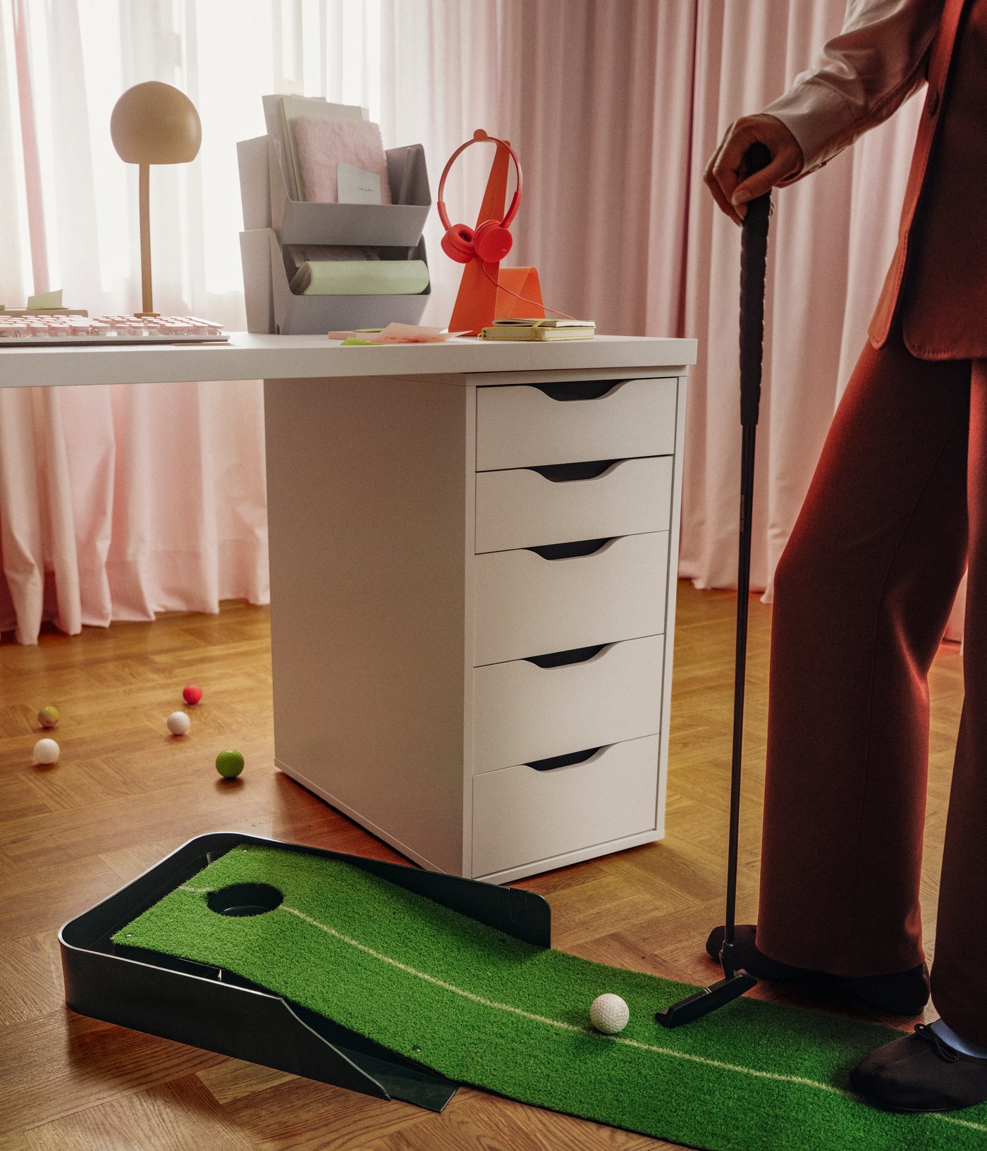 Una cassettiera ALEX bianca, dotata di eleganti maniglie a incavo, sostiene una scrivania bianca. Accanto, una persona si esercita nel minigolf.