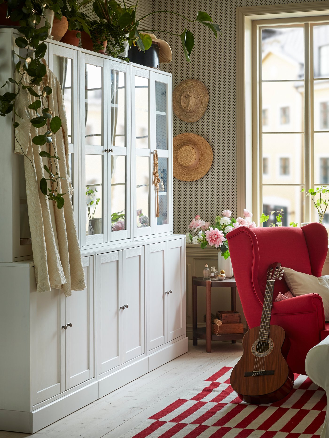 A white storage combination with glass doors in a red and white living room. Green plants are placed on top.