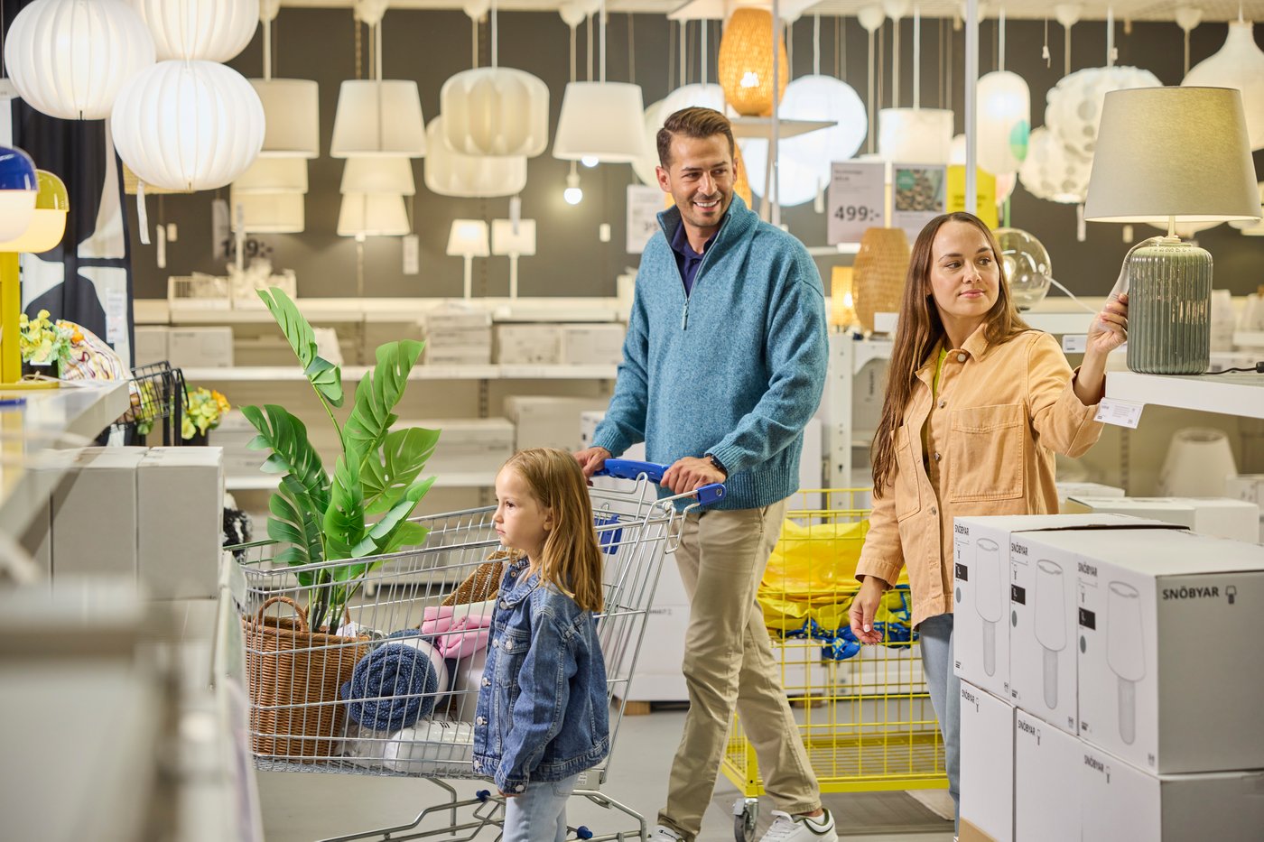 Eine Familie schiebt einen Einkaufswagen durch eine Lampenabteilung im IKEA Einrichtungshaus. Im Wagen sitzen ein Kind und eine Pflanze, während viele Leuchten im Hintergrund hängen.