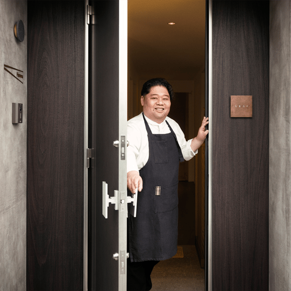 落ち着いたエントランスで、エプロン姿のスタッフがダークウッドのドアを開けて迎える高級レストラン入口
