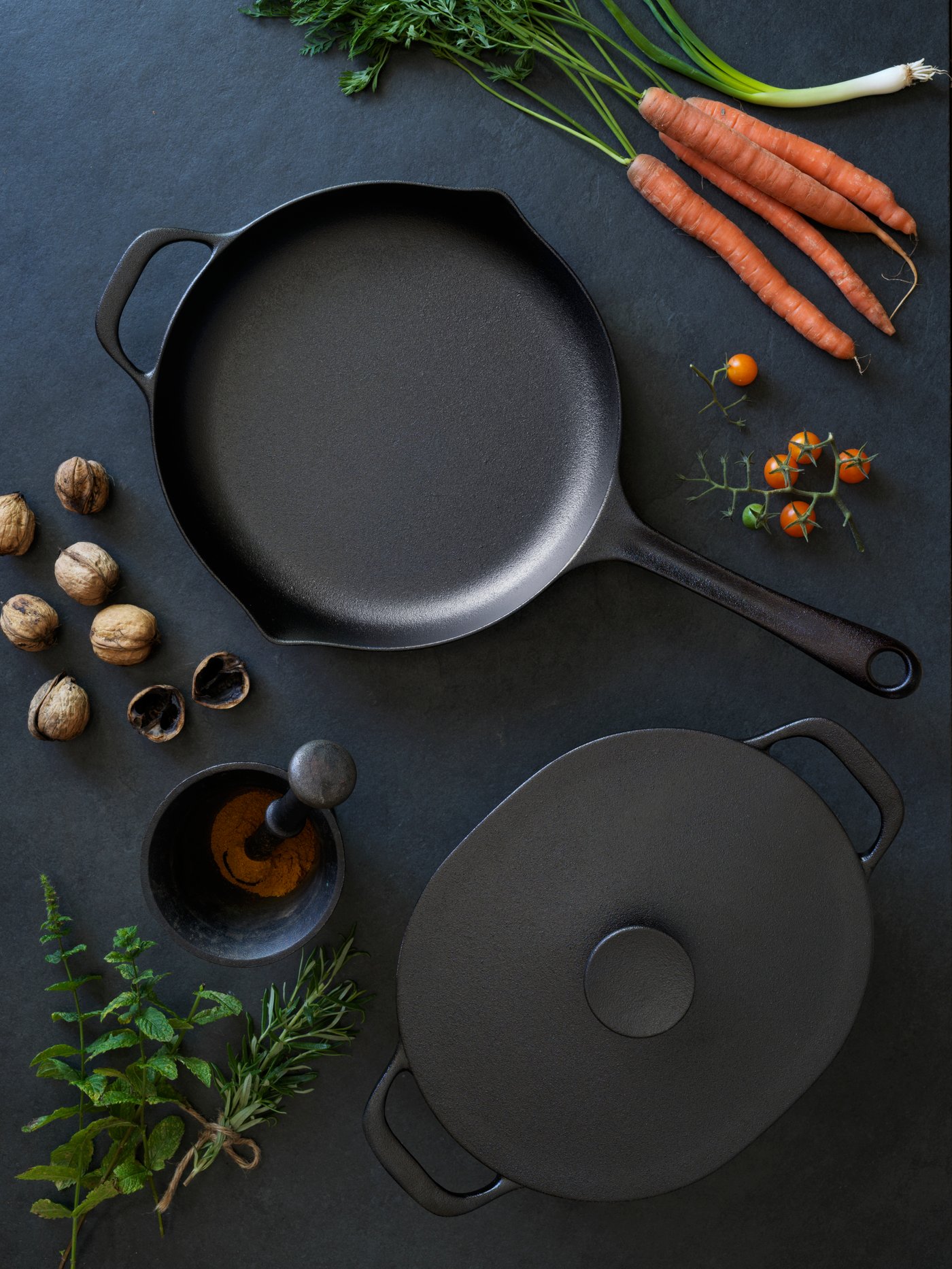 Una casseruola e una padella VARDAGEN su un tavolo in legno nero, circondate da verdure e altri ingredienti come carote e noci.