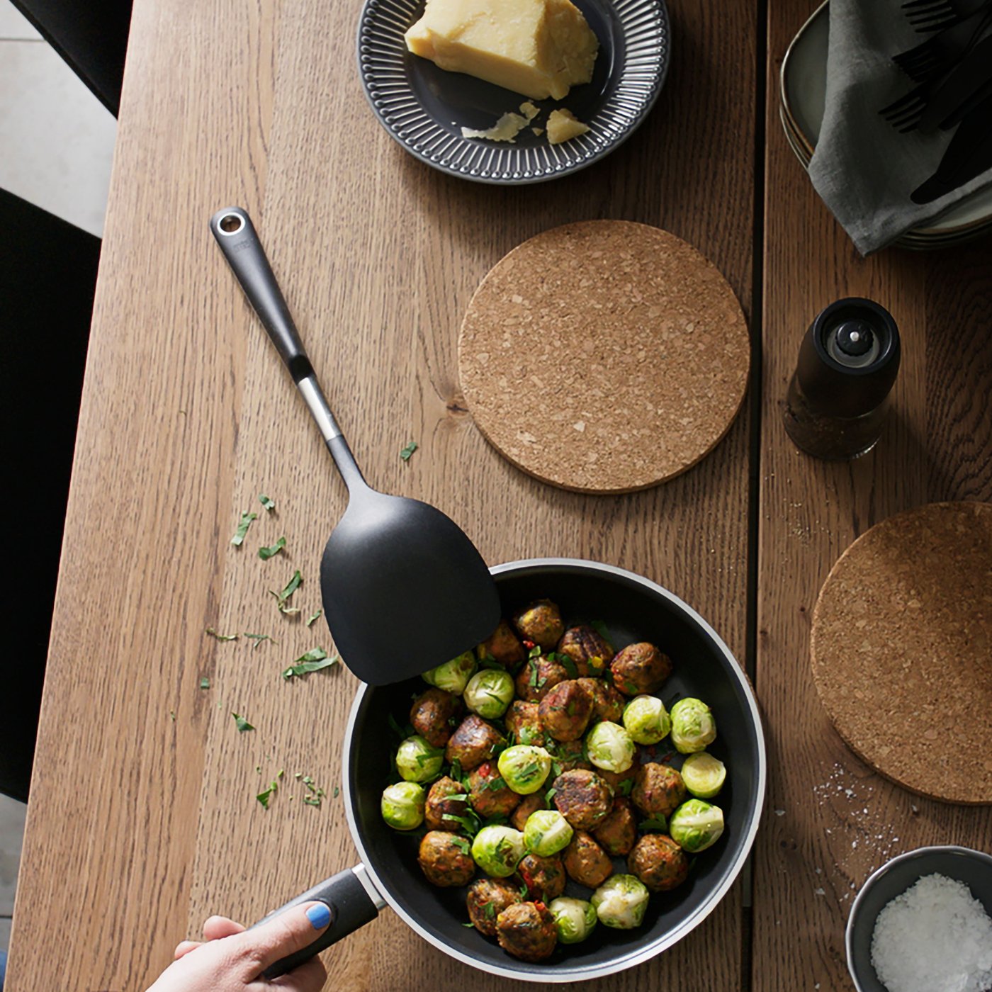 A table with a KAVALKAD frying pan in black containing brussels sprouts and HUVUDROLL plant balls, a HEAT pot stand in cork.