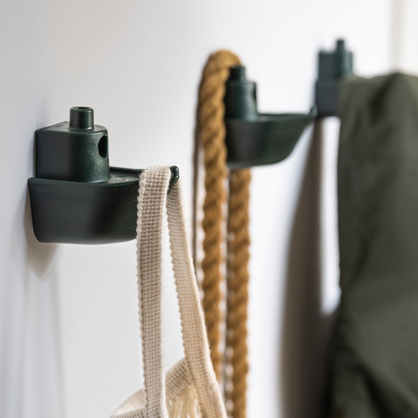 ثلاث خطافات KNOPARE خضراء مثبتة على حائط، اثنان منها يحملان أكياسًا بينما يحمل الثالث سترة.