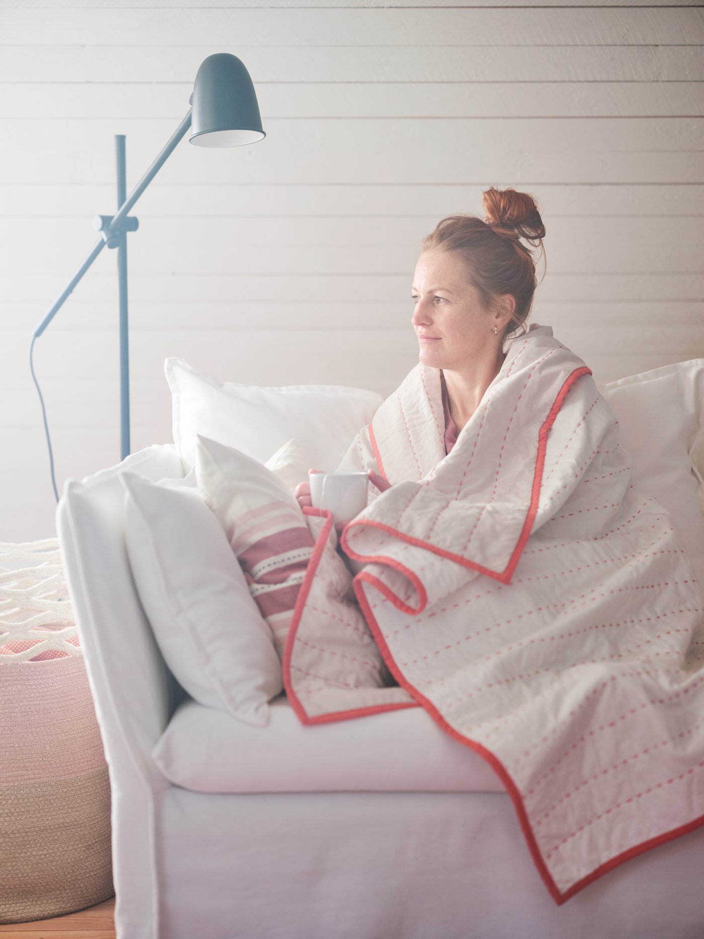 A woman is snuggled up with a cup of tea in a cotton VÅRDANDE throw on a white sofa.