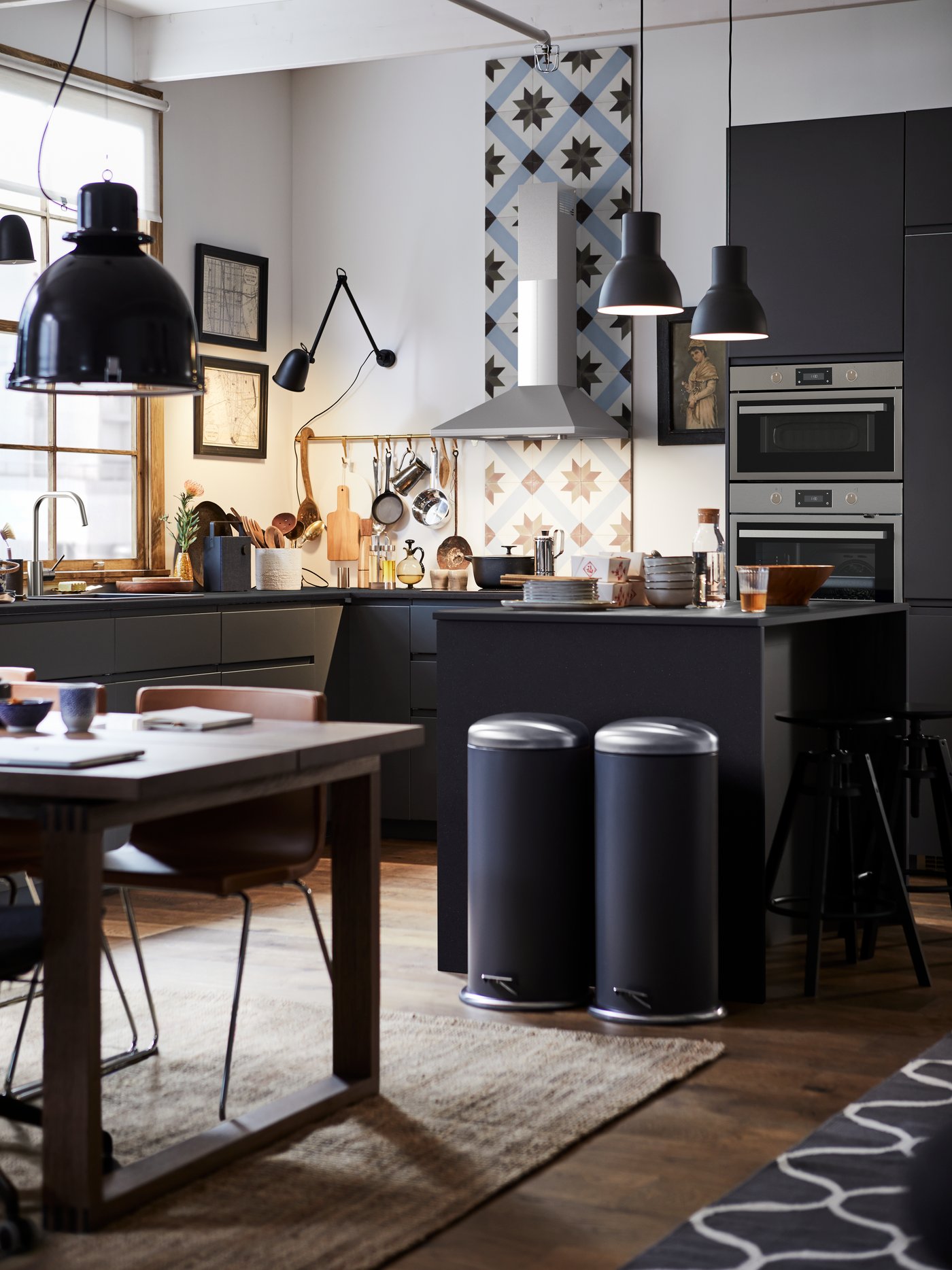Una cocina moderna con un armario bajo gris con cajones, una mesa, cubos con pedal, pantallas para lámparas de techo y una silla.