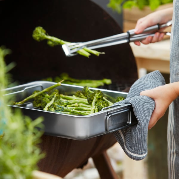 A KONCIS roasting pan filled with asparagus being held on the edge of a barbeque.