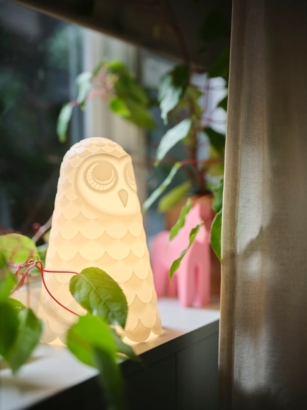 A white/owl SOLBO LED table lamp that is switched on sits on a window sill beside an off-white LENDA curtain with tie.