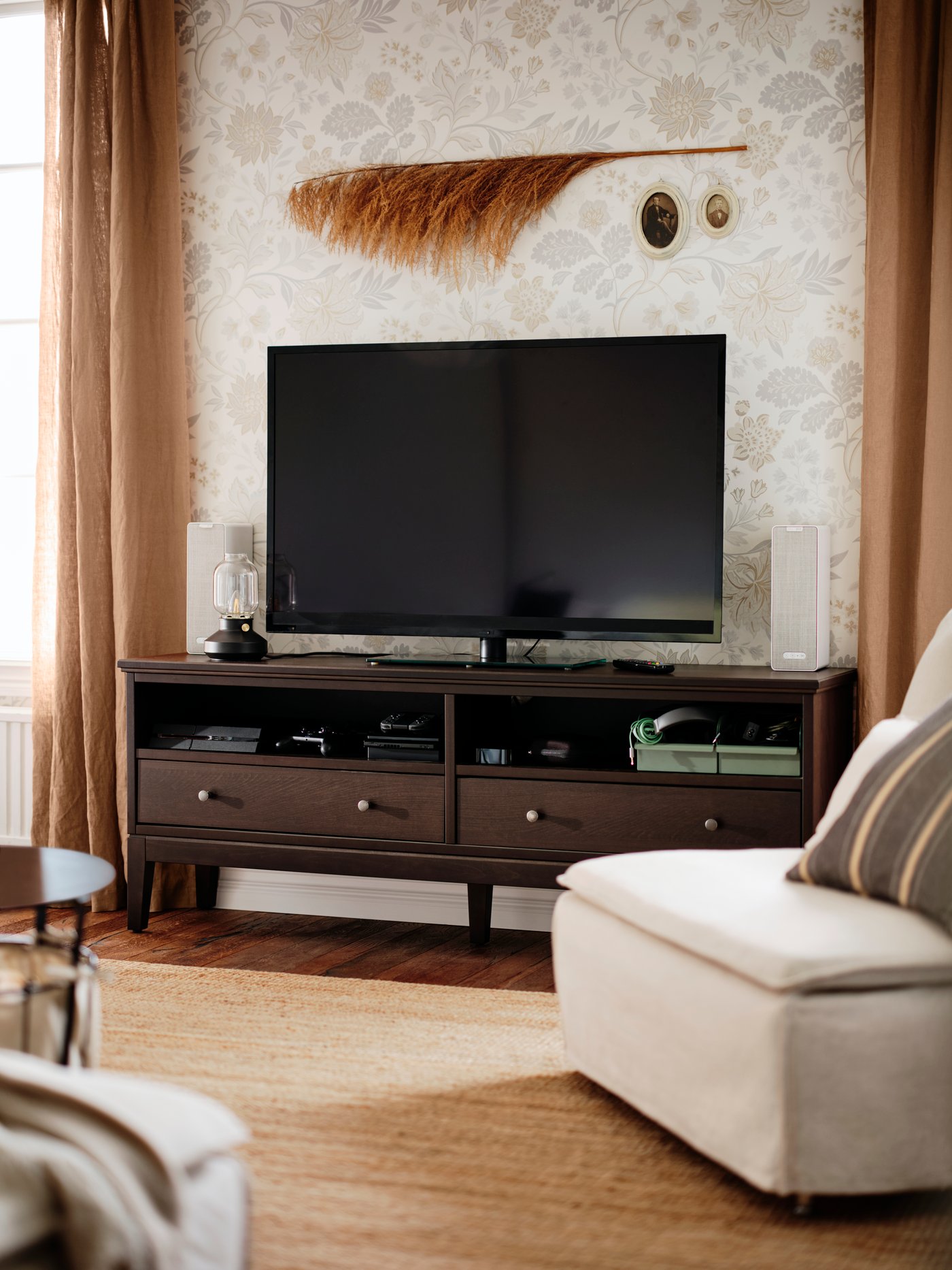 Un mueble para TV IDANÄS con un televisor entre dos parlantes encima. En la pared de arriba hay una pluma larga y dos cuadros ovalados.