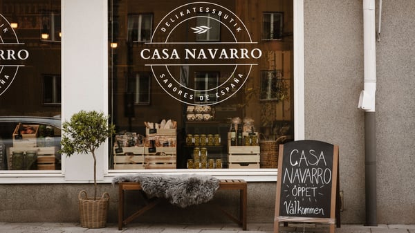 Texten 'Casa Navarro' på ett skyltfönster där man kan se in i en spansk delikatessbutik. Utanför fönstret står en NÄMMARÖ bänk med ett fårskinn på, ett olivträd och en skylt med texten 'Casa Navarro', 'Öppet', 'Välkommen'.
