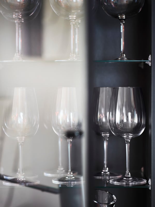 A number of STORSINT wine glasses stand on the glass shelves of a black/oak veneer BOASTAD high cabinet.