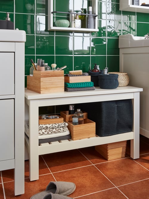 Bathroom with a white bench with a basket, round boxes, a bathroom set, and folded towels. A wall  shelf is on the wall. 