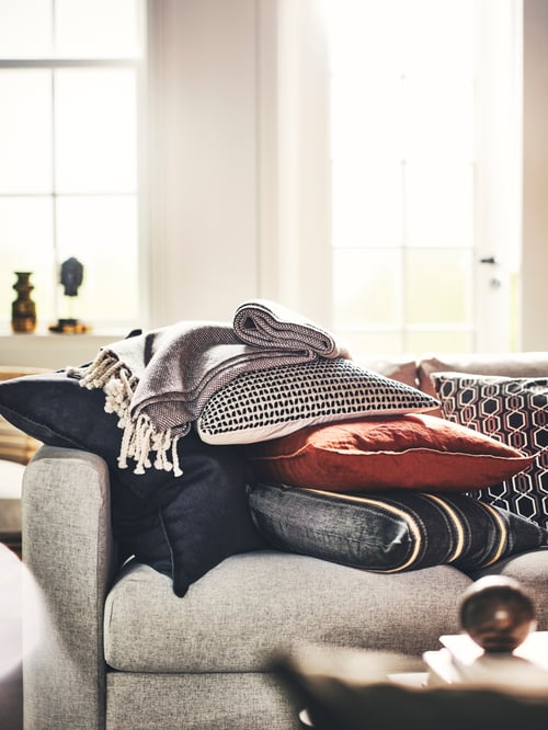 Nahaufnahme eines Kissenstapels auf einem Sofa mit einem Kissen in einem KUSTFLY Kissenbezug, einem DYTÅG Kissenbezug und einem ULLTISTEL Plaid.