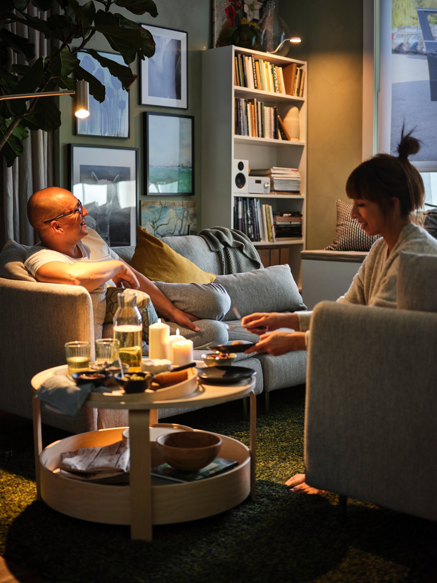 Two people are relaxing on an IKEA SÖDERHAMN sofa in a cosy living room with a round coffee table set with drinks and candles.
