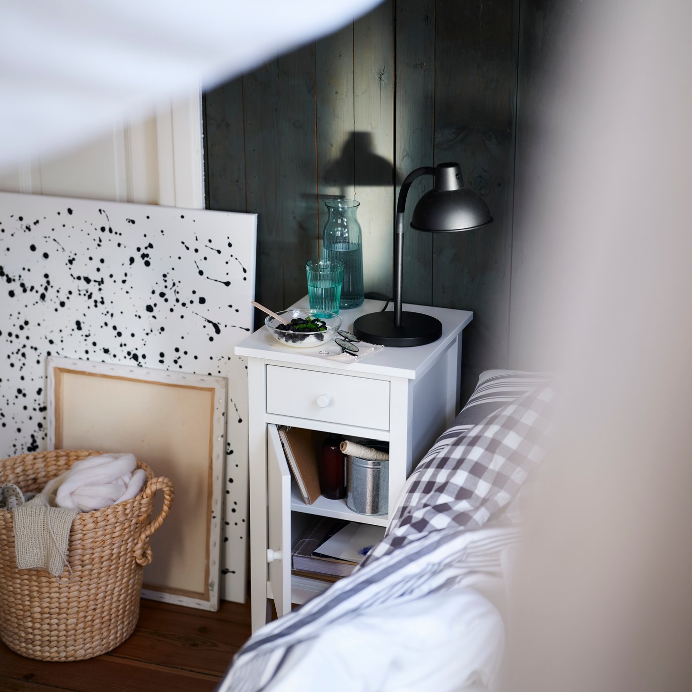 Szafka nocna GULLABERG w kolorze białym z szufladą i otwartą półką obok łóżka w sypialni z ciemnymi panelami.