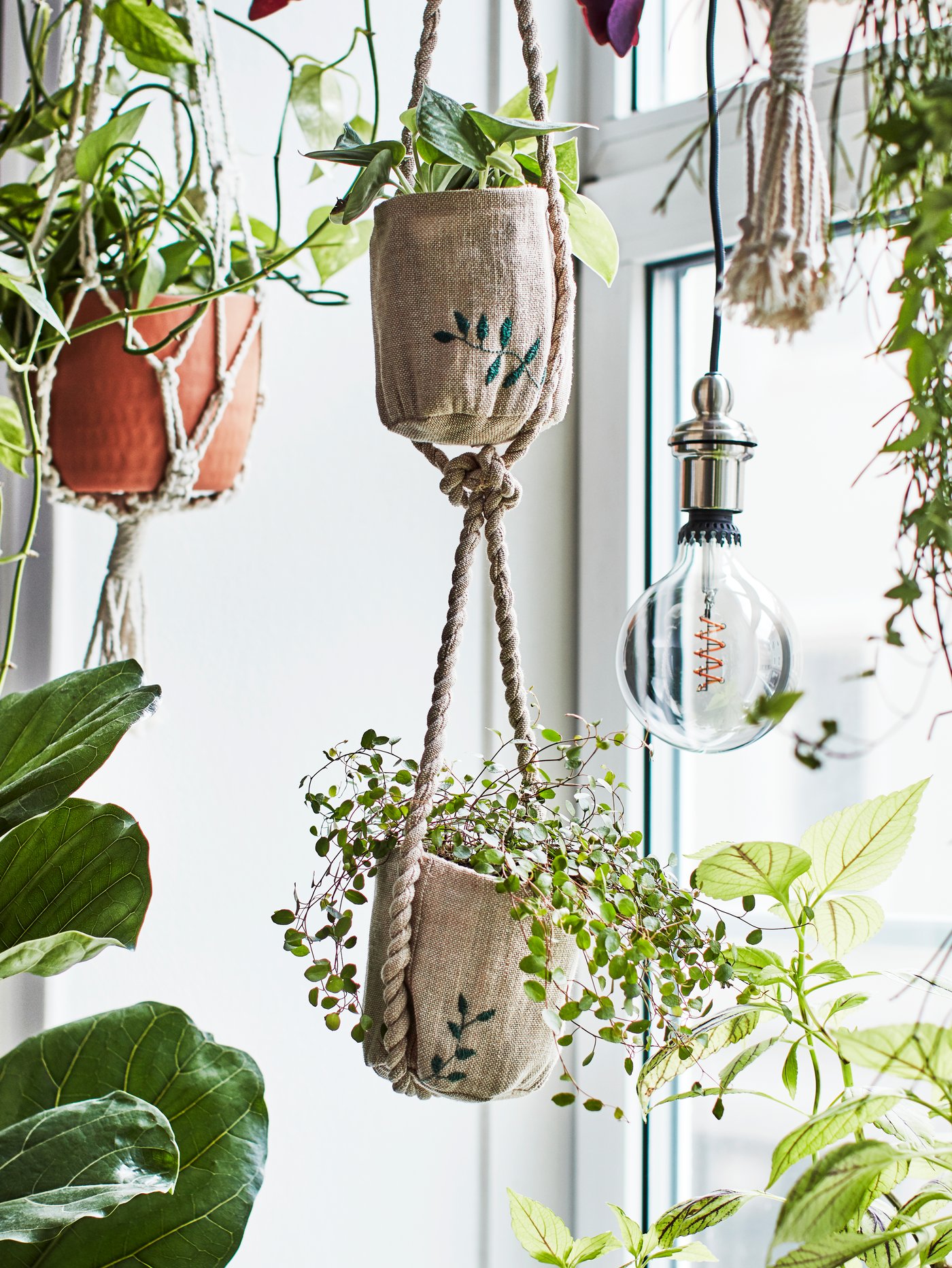 Handmade jute planters with embroidered details hanging at different heights in a window inside a home.