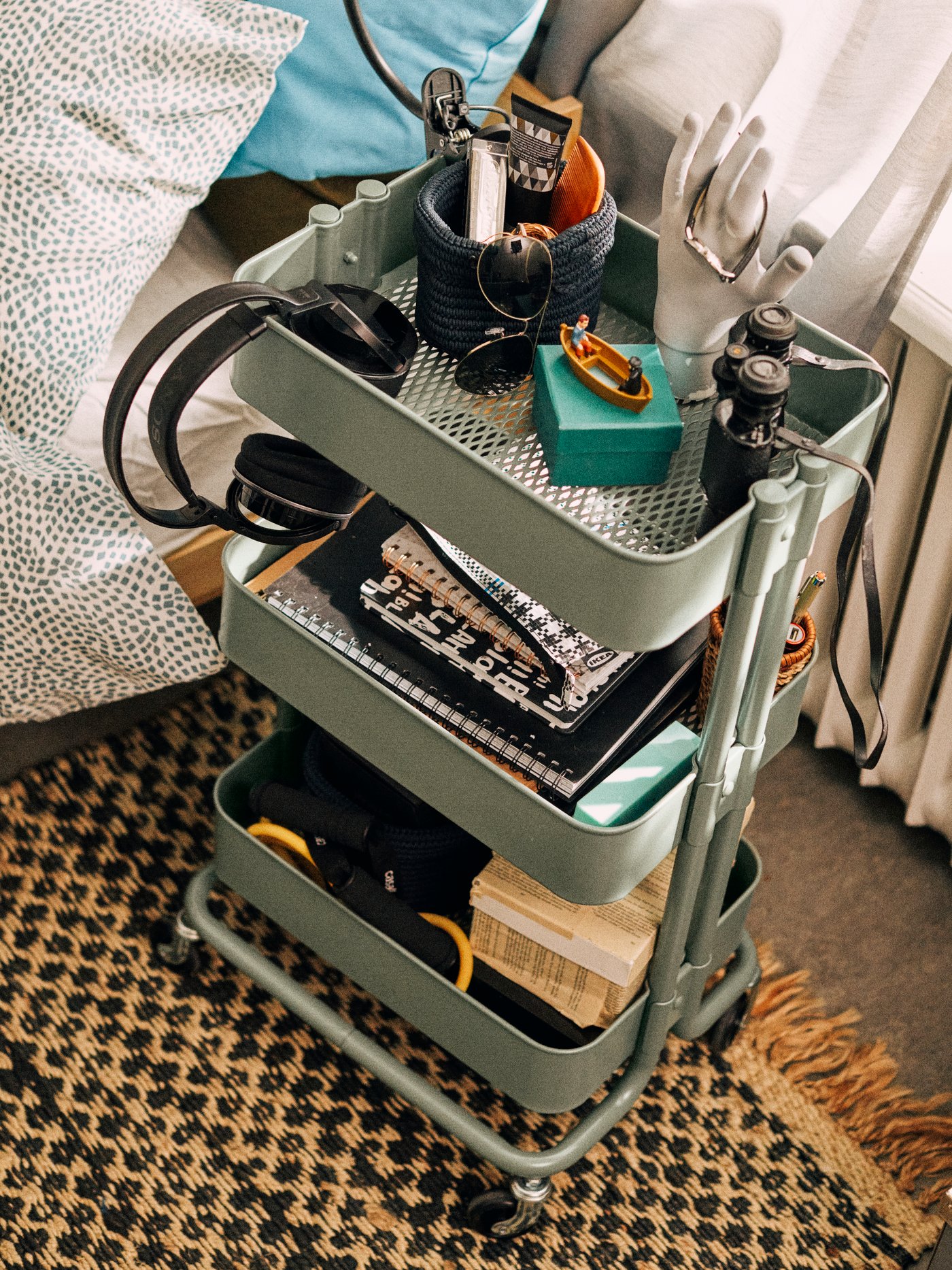 Carrito RÅSKOG en verde grisáceo sobre una esterilla junto a una cama. En el carrito hay cuadernos y otras cosas.