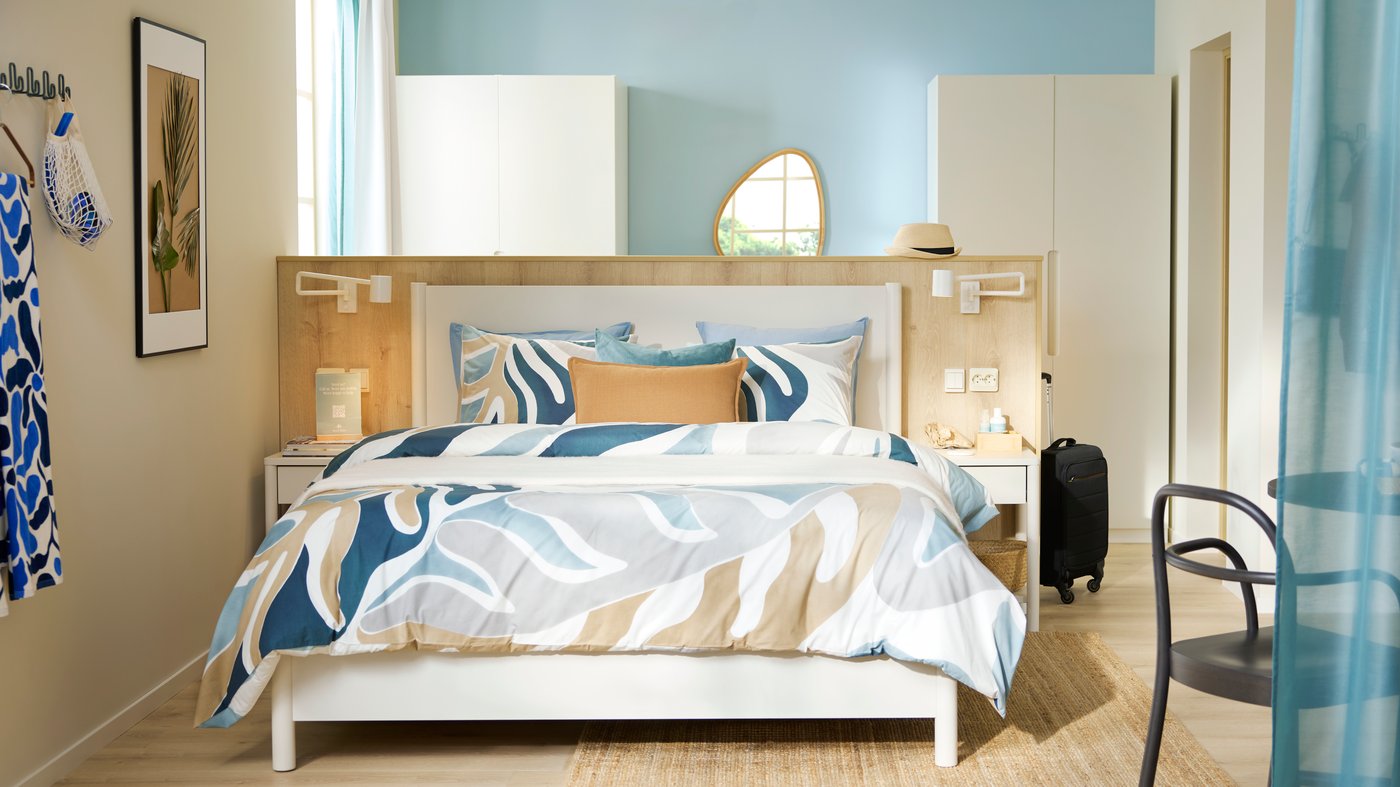 An airy, beachy-feeling hotel room has beige and light teal walls with an off-white TONSTAD bed frame in the room’s centre.
