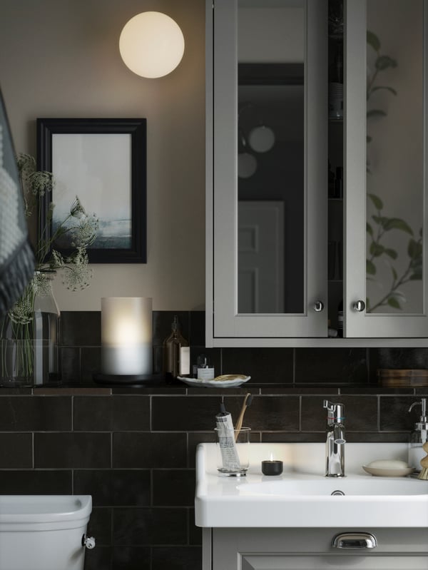 A bathroom with a light gray TÄNNFORSEN mirror cabinet with doors and a round FRIHULT lamp on the wall. 