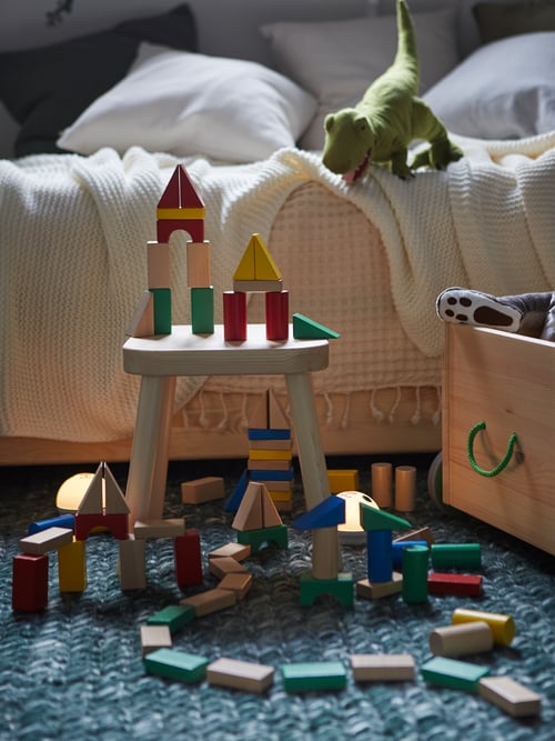 Multi-coloured pieces from UNDERHÅLLA wooden building block set are on a stool and on the floor of a child’s bedroom.
