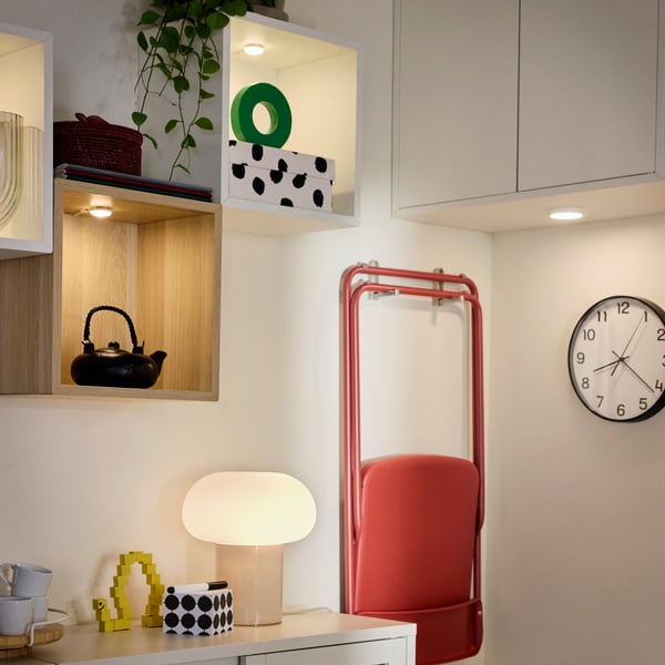 A room with a VIHALS foldable chair in red, folded and hanging on the wall, a clock on the wall and a DEJSA table lamp.