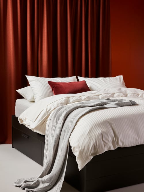 Bedroom with a bed frame with storage with duvet cover and multiple pillows, with a red pair of curtains in the background.