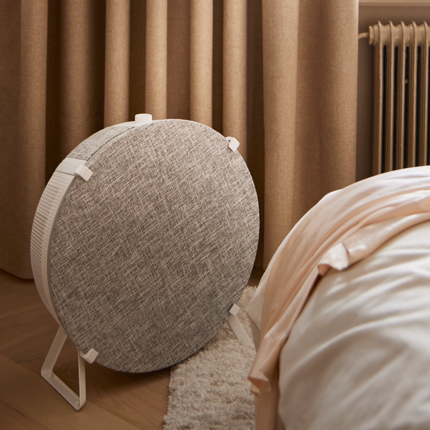En rund, tygklädd högtalare står på golvet bredvid en säng med textilier i beige toner. I bakgrunden syns bruna gardiner och en fönsterbräda med böcker.