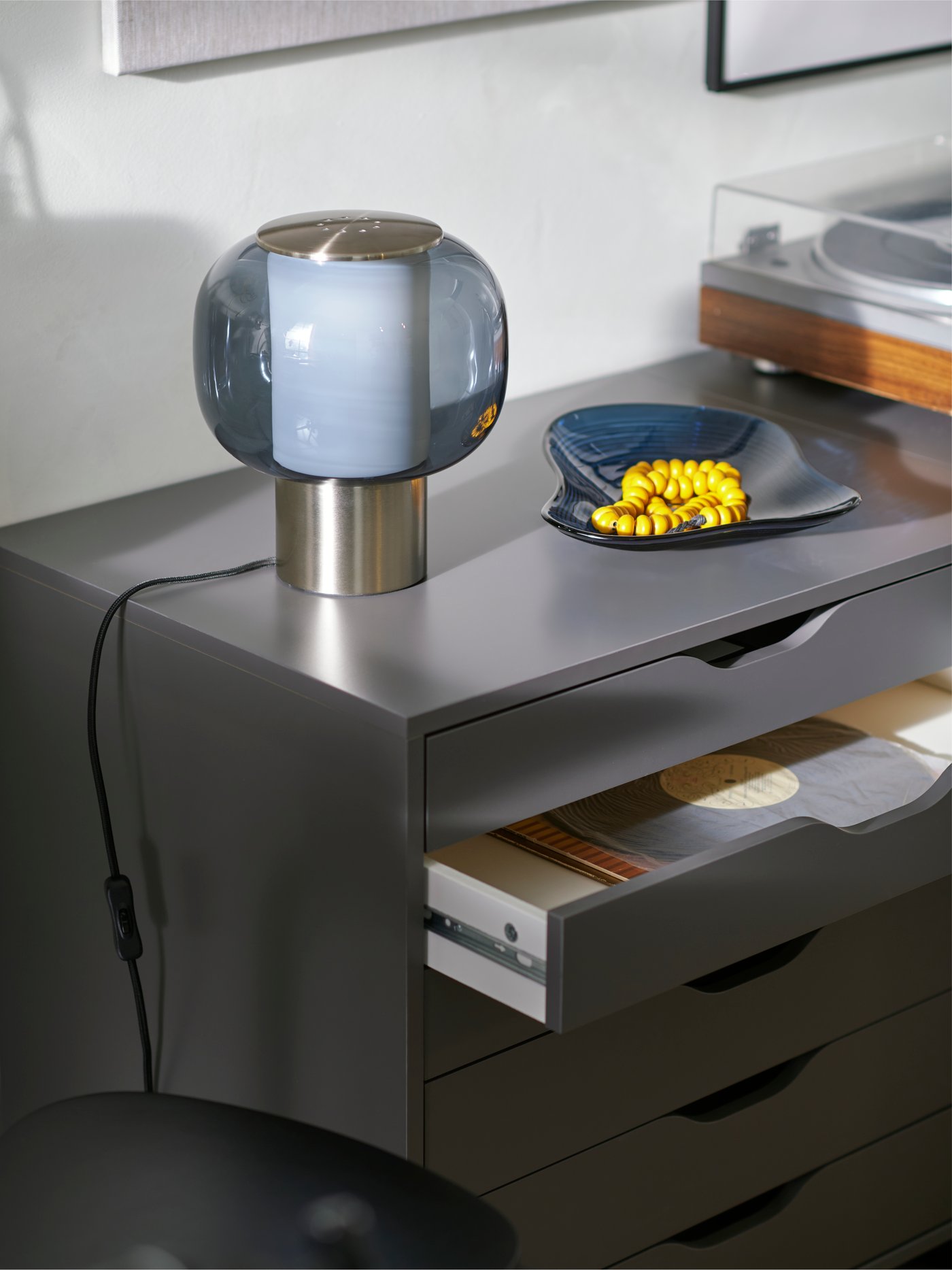 Eine HÖGVIND Tischleuchte und eine blaue HÄCKPOPPEL dekorativ Schüssel mit einer gelben Holzkette stehen auf einer dunkelgrauen ALEX Schublade Einheit.